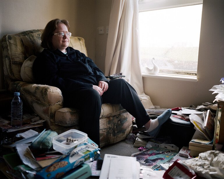 A woman sits in her lounge surrounded by papers and magazines