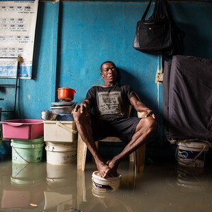 A man sits in his room with his feet up on a paint bucket to avoid the floodwater