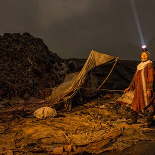 A midwife stands in a landfill site at night, searching for things to sell