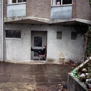 A man sits on a chair in the doorway of his flat. 