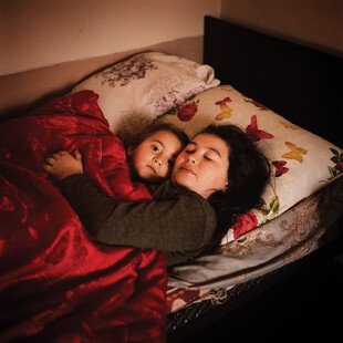 Two girls lying in a bed wrap their arms around each other.
