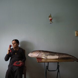 A woman holds a telephone to her ear, a big fish on the table beside her.