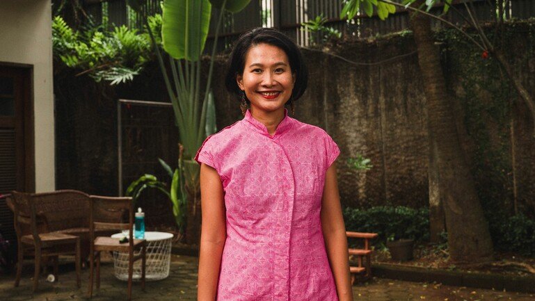 An image of a woman standing up, seen from the hip up, and wearing a pink shirt. She is at centre frame, smiling to the camera, with plants behind her.